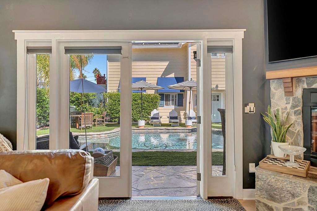 The image showcases a living room with a view of a backyard pool area through open sliding glass doors. The interior features a stone fireplace and a glimpse of a leather sofa, while the exterior view includes a pool, patio furniture, and a well-maintained lawn, creating an inviting indoor-outdoor living space. The scene suggests a relaxed and luxurious lifestyle.