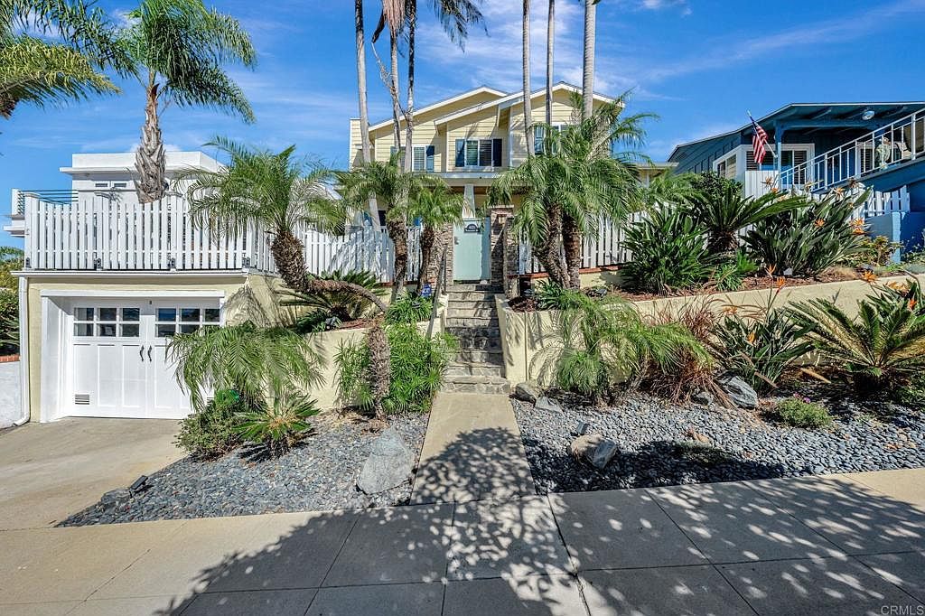 This is a front view of a two-story house with a yellow exterior and a white garage door. The property features a landscaped front yard with palm trees, various plants, and decorative rocks. A set of stairs leads to the front door, and a white picket fence partially encloses the property.