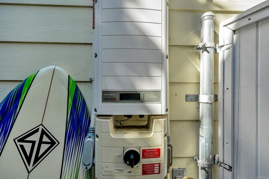 The image showcases a SolarEdge inverter mounted on the exterior wall of a house, positioned next to a surfboard. The inverter, with its visible on/off switch and warning labels, suggests a modern solar energy system. The surfboard adds a touch of coastal lifestyle, implying a connection to outdoor activities and a relaxed living environment.