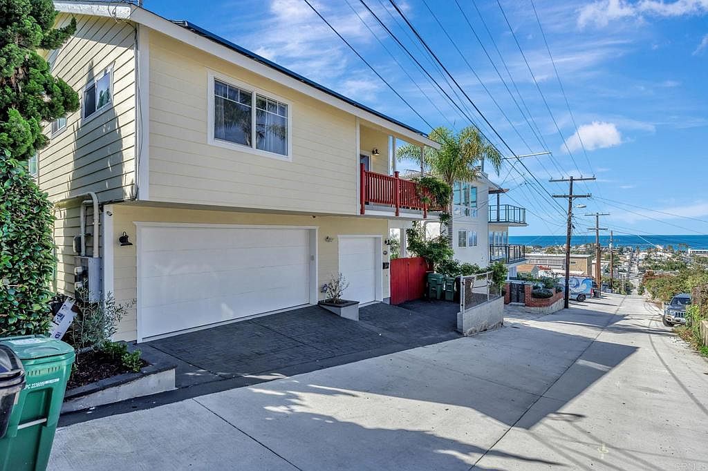 This is a front exterior view of a two-story house with a light yellow facade and white trim. The house features a large two-car garage, a smaller single-car garage, and a red-railed balcony. The property is situated on a sloped street with a view of the ocean in the distance, creating a desirable coastal living impression.
