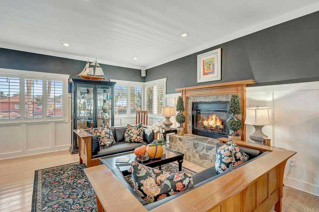 This is an interior shot of a living room featuring a stone fireplace with a wooden mantel, flanked by built-in seating with floral cushions. The room has a dark gray accent wall above white wainscoting, and a large cabinet displays a model ship. The space is well-lit with natural light from the windows and recessed lighting, creating a cozy and inviting atmosphere.