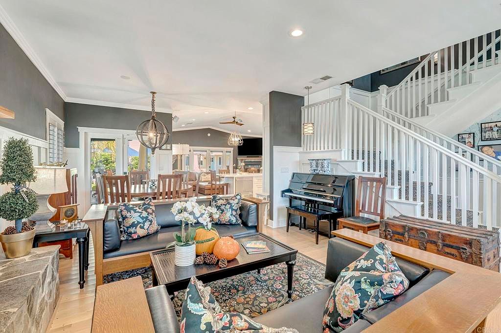 This is an interior shot of a living room that flows into a dining area and kitchen. The living room features two sofas with decorative pillows, a coffee table with pumpkins, and a fireplace. A piano sits near a staircase with white railings. The overall style is traditional with a touch of modern flair.