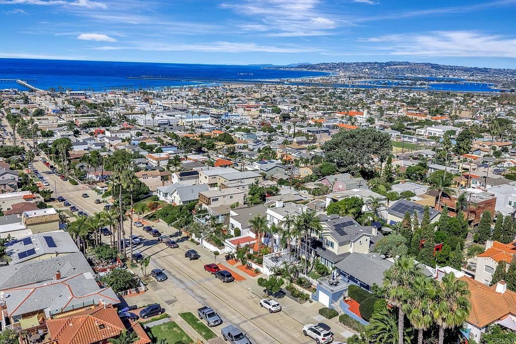 This aerial view showcases a dense residential neighborhood near the ocean. The houses feature a mix of architectural styles with many having solar panels. Palm trees line the streets, adding a touch of coastal charm, and the ocean is visible in the background, suggesting a desirable location.