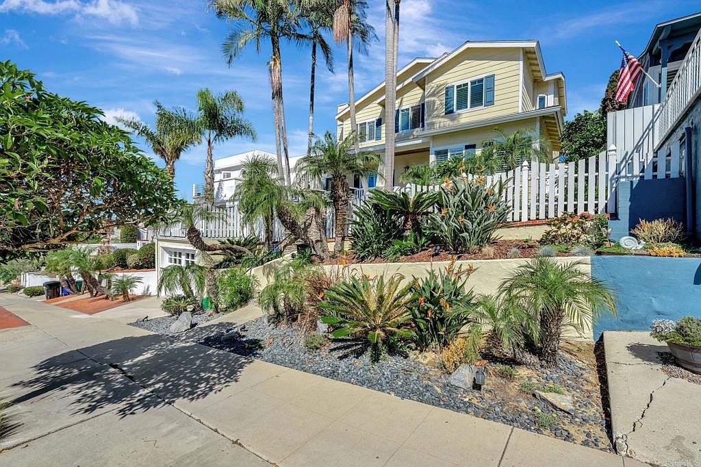 This is a front view of a two-story yellow house with white trim and a white picket fence. The property is landscaped with palm trees, various plants, and decorative rocks. The house is situated on a slight hill, adding to its curb appeal.