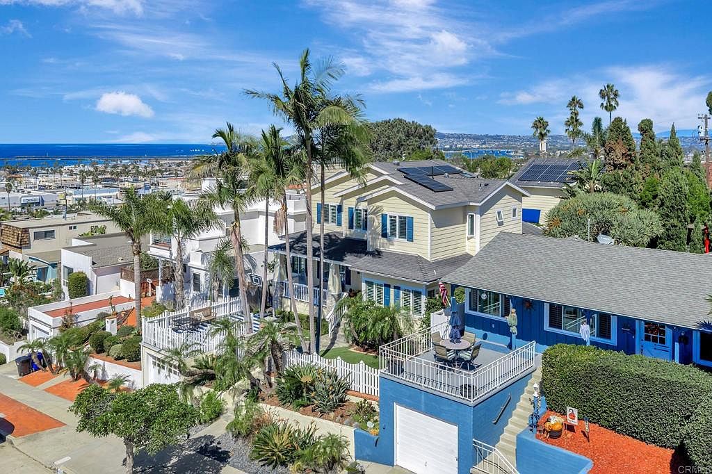 This aerial view showcases a collection of charming coastal homes, with a clear view of the ocean in the background. The houses feature a mix of architectural styles, including a yellow two-story house with blue shutters and a blue house with a rooftop deck, all surrounded by lush greenery and palm trees. Solar panels are visible on the roofs, adding a modern touch.