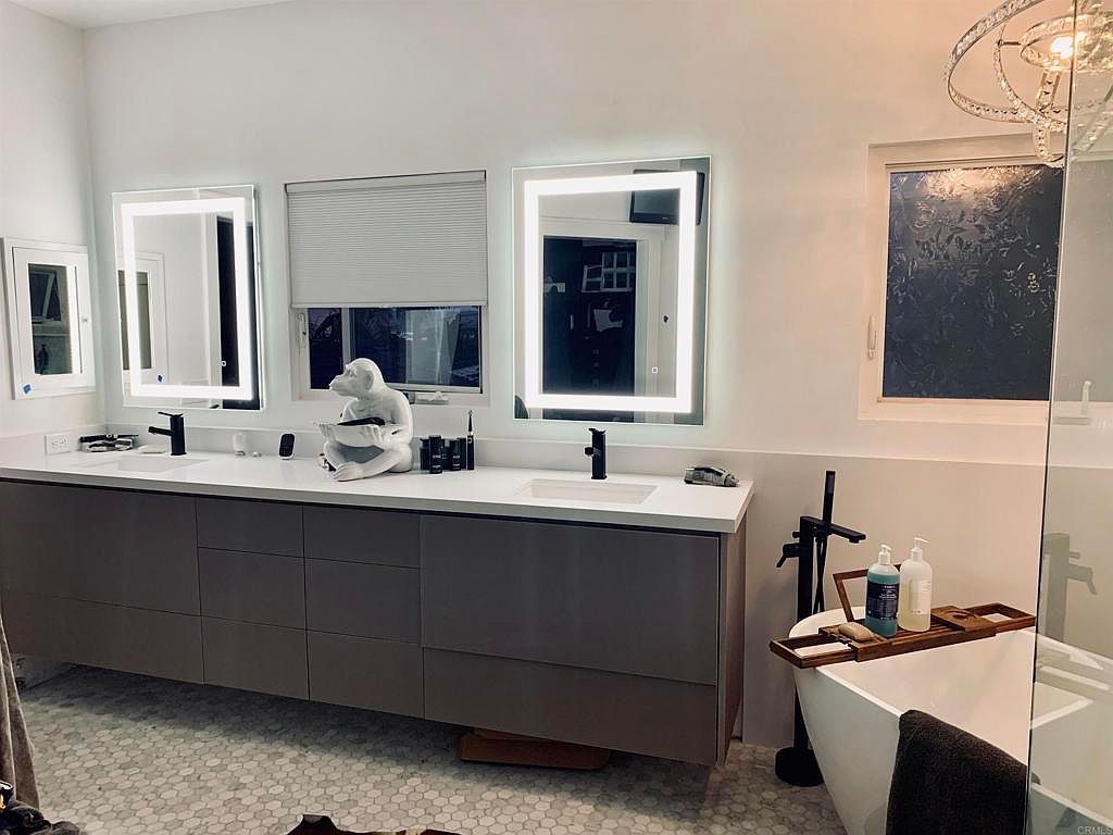 This is a modern primary bathroom featuring a long double vanity with gray cabinets and a white countertop. Two illuminated mirrors hang above the sinks, and a white sculpture of a monkey sits on the counter. A freestanding bathtub with black fixtures is positioned near a window with privacy film, and the floor is covered in a small hexagonal tile pattern.