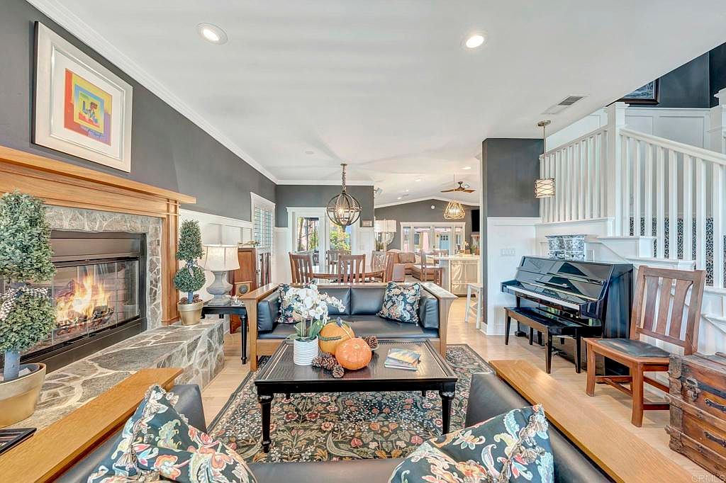 This is an interior shot of a living room featuring a stone fireplace with a wooden mantel, a black leather sofa with floral pillows, and a black coffee table with pumpkins and books. The room has hardwood floors, gray walls, and a staircase with white railings. The style is traditional with a touch of modern.