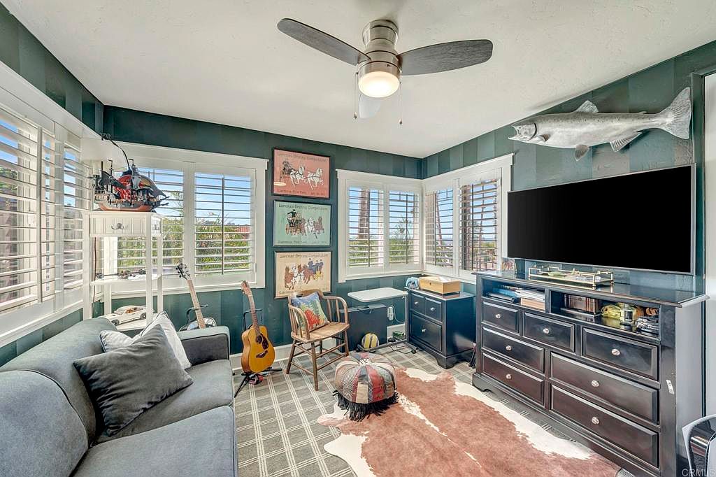 This is an interior shot of a bedroom featuring a gray sofa with pillows, a guitar, and a mounted fish above a dark wood dresser with a television. The room has a plaid rug with a cowhide rug on top, and the walls are a dark green with white trim. The room has multiple windows with white shutters, and the overall style is eclectic and cozy.