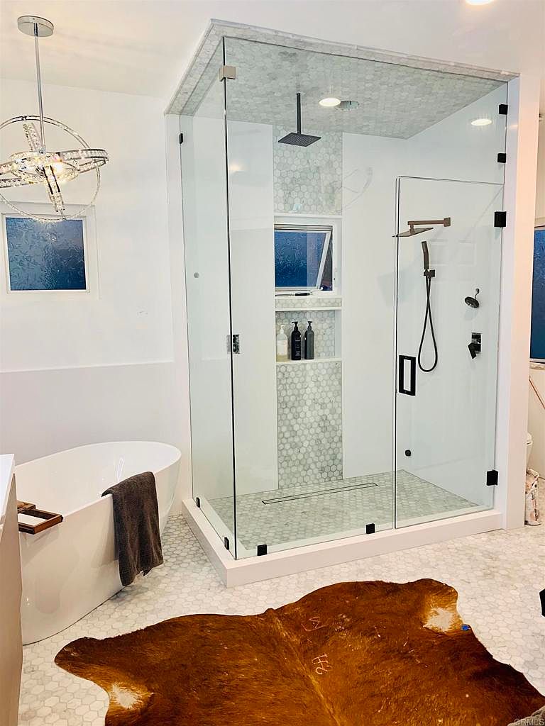 This is a luxurious primary bathroom featuring a freestanding white bathtub with a dark towel draped over the side. A glass-enclosed shower with a rainfall showerhead and built-in niche is the focal point, complemented by hexagonal tile flooring and a modern chandelier. A brown cowhide rug adds a touch of rustic elegance to the space.