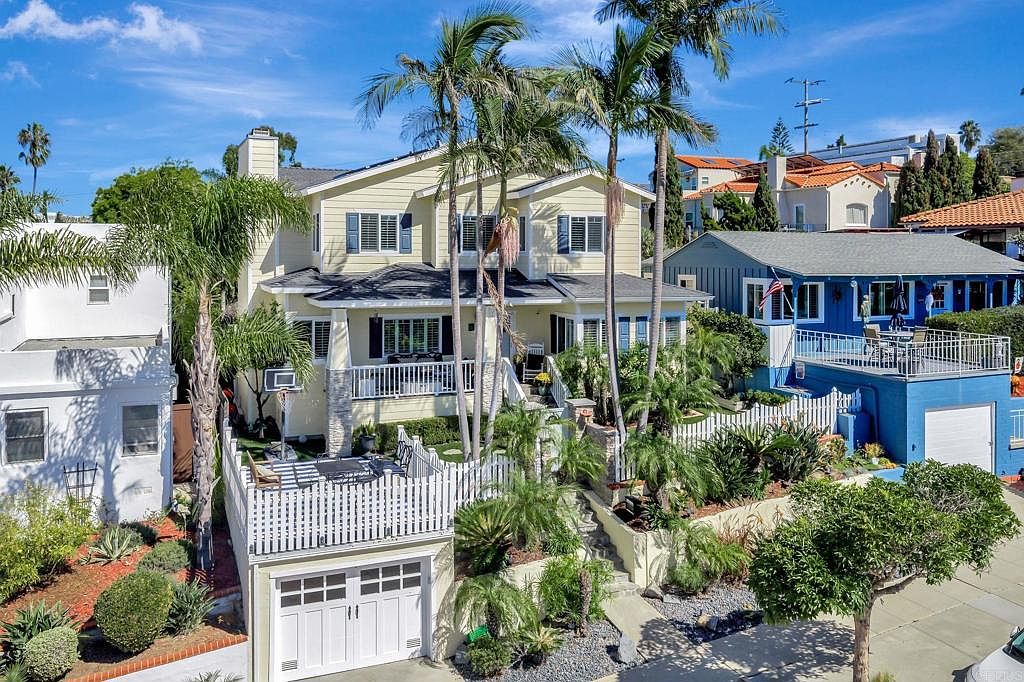 This is a front exterior view of a multi-story house with a well-maintained yard. The house features a light yellow exterior with dark shutters and a dark roof. The property includes a garage with a white picket fence above, lush landscaping, and tall palm trees, creating a welcoming and upscale curb appeal.