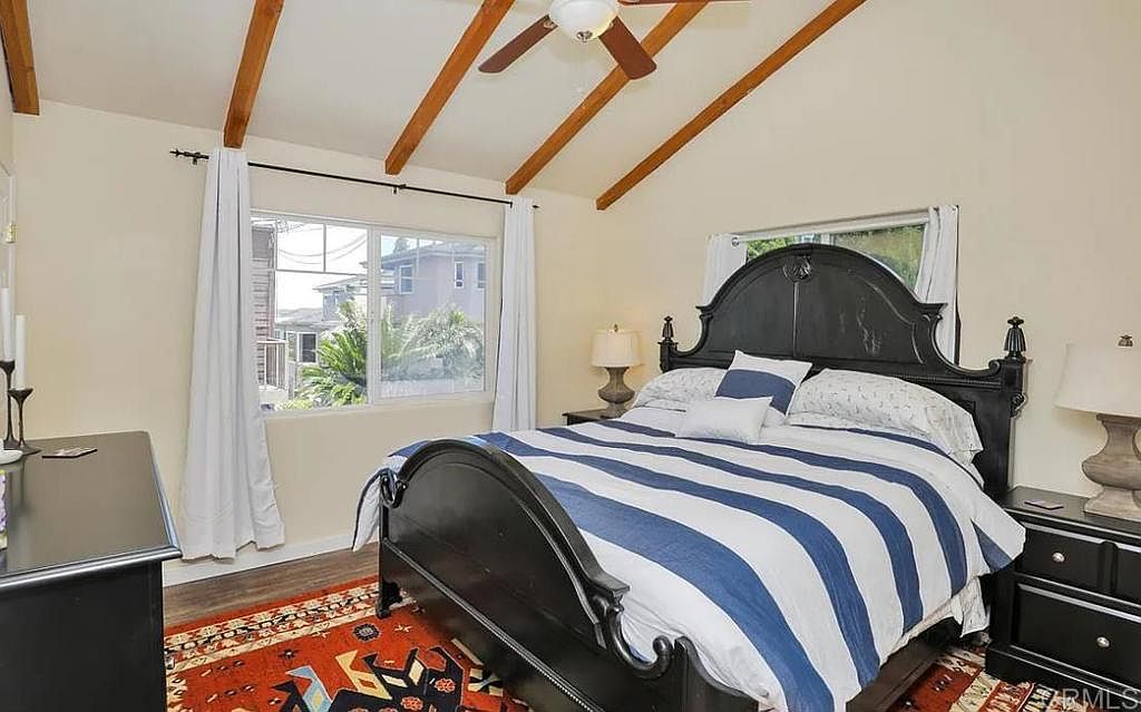 This is a primary bedroom featuring a black wooden bed frame with a blue and white striped comforter. The room has exposed wooden beams on the ceiling and a window that provides natural light. There is a nightstand with a lamp on either side of the bed, and a rug on the floor.
