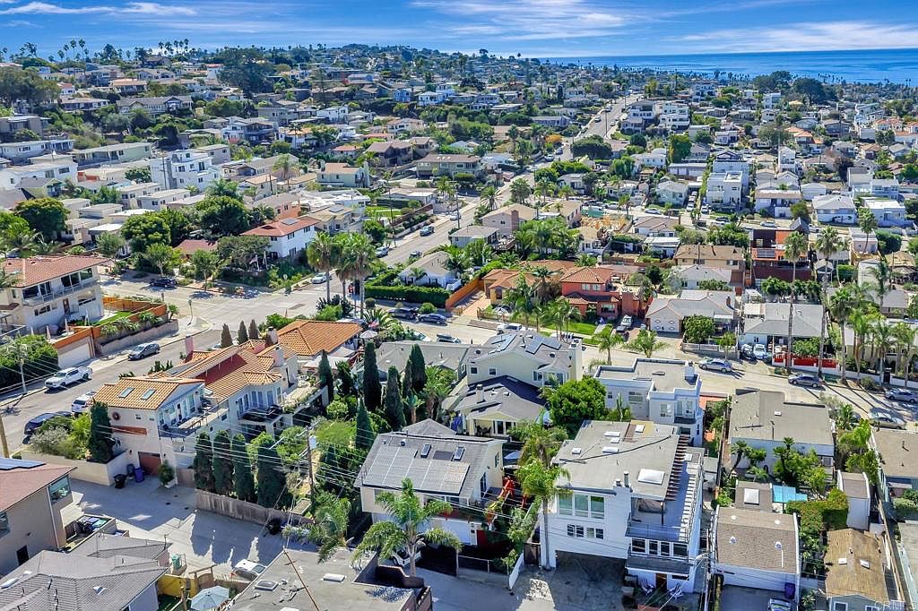 This aerial shot showcases a dense residential neighborhood with a mix of single-family homes, many featuring red tile roofs and solar panels. Lush greenery and palm trees are interspersed throughout, leading towards a distant view of the ocean under a partly cloudy sky. The perspective gives a comprehensive view of the community's layout and proximity to the coast.
