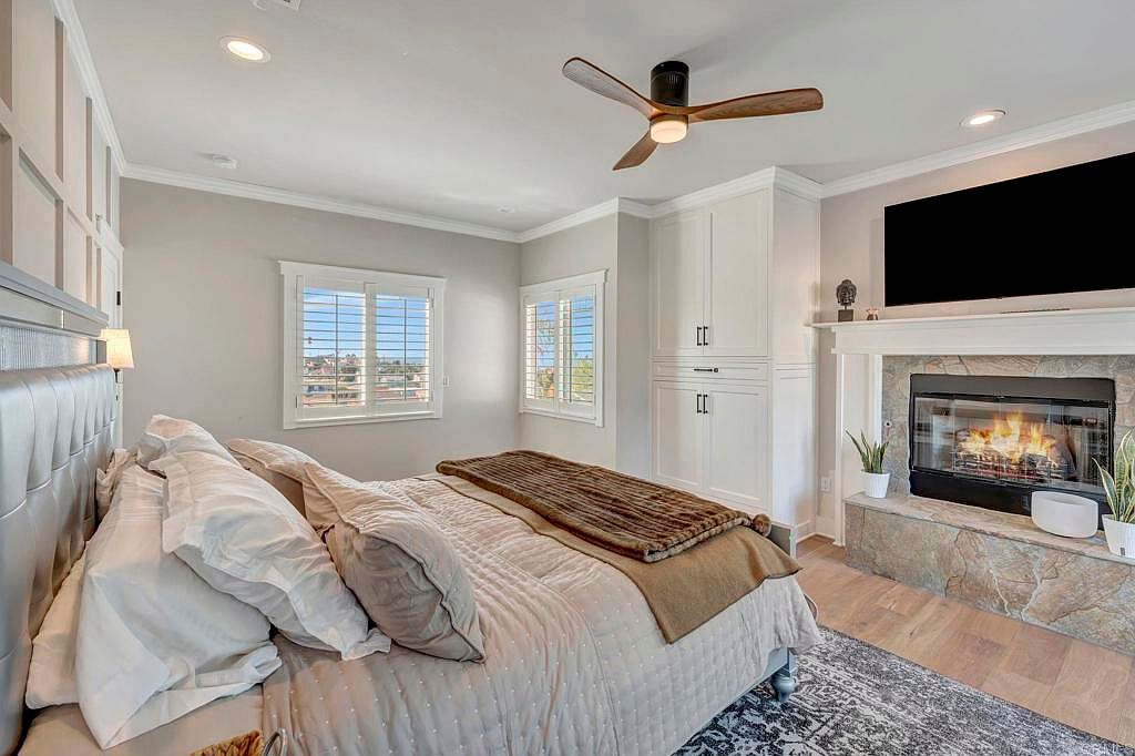 This is a primary bedroom featuring a large bed with multiple pillows and a brown throw blanket. The room has two windows with white shutters, built-in white cabinets, and a fireplace with a mounted television above it. The flooring is hardwood, and there is a ceiling fan.