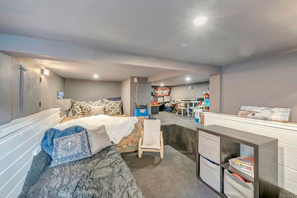 This is a view of a finished basement area, featuring a multi-level design with sleeping areas and a play space. The room is painted in neutral tones, and the lower level includes a bed with decorative pillows and a small chair. A storage unit with fabric bins is visible, and the upper level appears to be a play area with a table and various items.