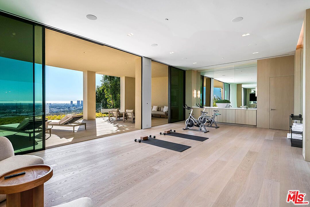 This expansive, modern home gym features light hardwood flooring and floor-to-ceiling glass sliding doors that open onto a spacious balcony with panoramic city views. The room is equipped with two stationary exercise bikes, yoga mats, and a sleek built-in vanity area with a large mirror, creating a bright and airy atmosphere. The minimalist design and seamless indoor-outdoor transition emphasize a luxurious, wellness-focused lifestyle.