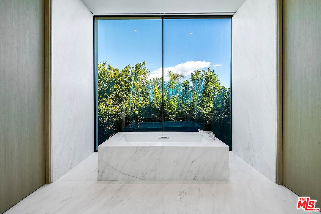 This luxurious primary bathroom features a minimalist, spa-like aesthetic centered around a large, marble-clad soaking tub. The room is framed by floor-to-ceiling marble walls and a massive window that provides an unobstructed, serene view of lush green trees. The clean lines and high-end materials create an atmosphere of sophisticated tranquility and modern elegance.