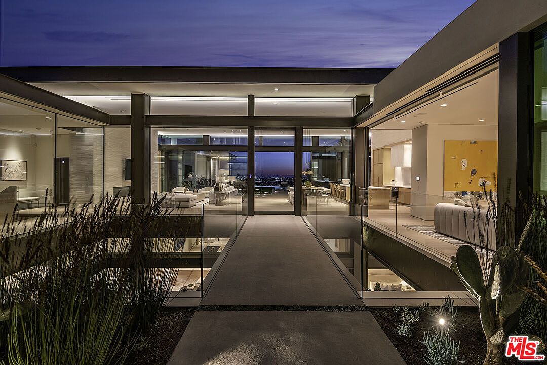 This striking modern entryway features a sleek, elevated stone walkway that bridges over a lower level, leading directly into a grand, glass-walled living space. The architectural design emphasizes clean lines, expansive floor-to-ceiling windows, and a seamless transition between indoor and outdoor environments. The twilight setting highlights the warm interior lighting, creating an inviting and sophisticated atmosphere that showcases the home's contemporary luxury.