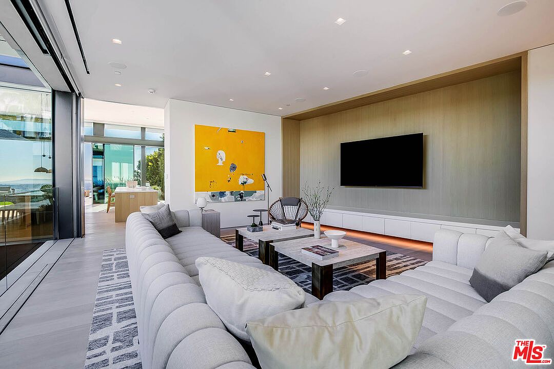 This sophisticated, modern living room features a large, plush sectional sofa arranged around two matching coffee tables, creating an inviting space for relaxation. A striking, oversized yellow abstract painting serves as a bold focal point against the clean white walls, while a wall-mounted television is integrated into a sleek, wood-paneled feature wall. The room is bathed in natural light from floor-to-ceiling glass doors that open to a scenic outdoor view, emphasizing an airy, open-concept design.