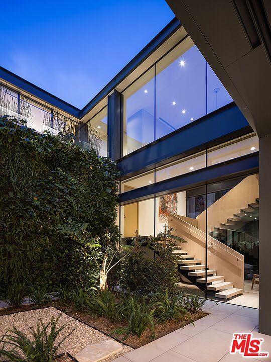 This striking architectural shot captures a modern, multi-level courtyard featuring a lush vertical garden wall and a minimalist gravel bed with ferns. A dramatic glass-enclosed staircase is visible through the floor-to-ceiling windows, seamlessly blending indoor and outdoor living spaces. The twilight lighting enhances the sophisticated, contemporary aesthetic of this high-end residential property.