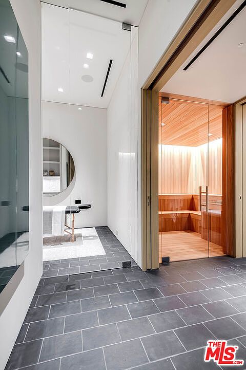 This luxurious spa-inspired space features a sleek massage room with a circular mirror and a dedicated sauna enclosed in glass and warm wood paneling. The room is finished with dark, large-format slate floor tiles that create a sophisticated contrast against the bright, minimalist white walls. The perspective captures a seamless transition between the treatment area and the sauna, emphasizing a high-end, wellness-focused lifestyle.