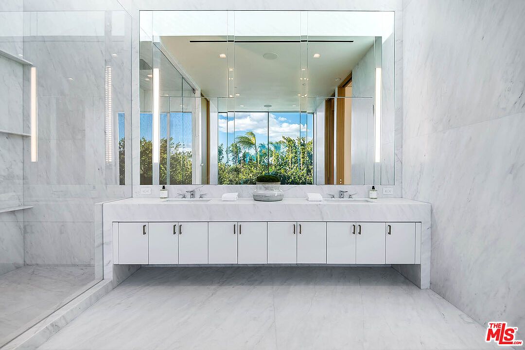 This luxurious primary bathroom features a sleek, minimalist design with floor-to-ceiling white marble tiling and a double vanity. A large, frameless mirror spans the wall, reflecting the bright natural light and lush greenery visible through the window. The space is characterized by clean lines, modern fixtures, and a bright, airy atmosphere that exudes high-end sophistication.