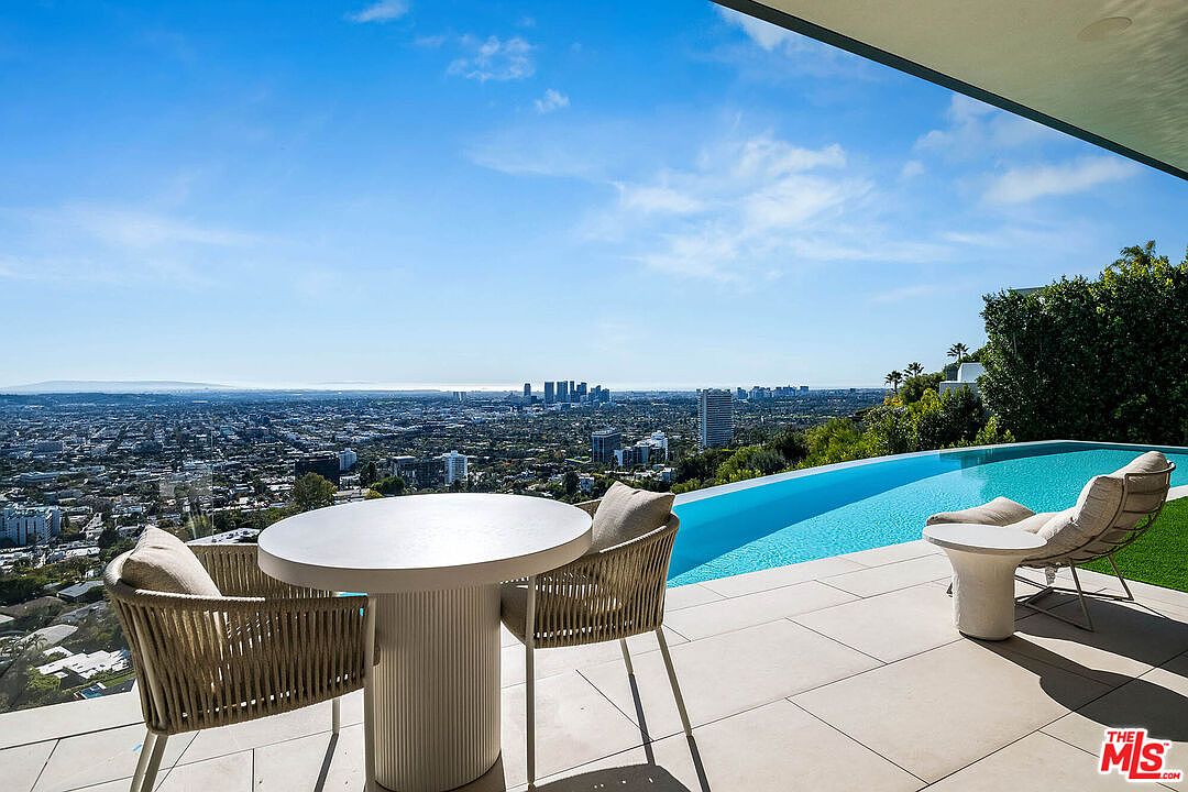 This stunning outdoor terrace features a sleek, modern infinity-edge pool that seamlessly blends into a panoramic view of the Los Angeles cityscape. The space is furnished with contemporary woven chairs and a minimalist round table, creating an inviting area for relaxation while overlooking the sprawling urban horizon. The clean lines and neutral stone flooring emphasize a luxurious, high-end architectural aesthetic.