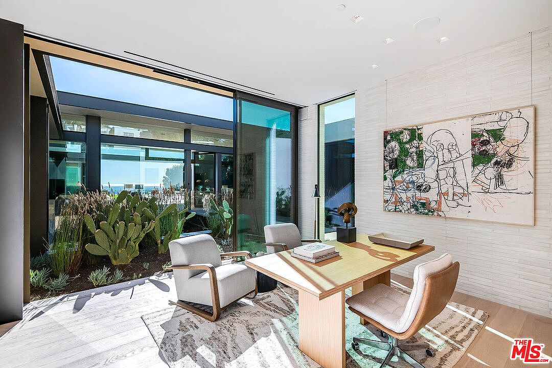 This sophisticated home office features a minimalist design with a light wood desk, two modern armchairs, and a large abstract art piece mounted on a textured white brick wall. Floor-to-ceiling glass sliding doors provide a seamless transition to an outdoor courtyard filled with lush cacti and greenery, creating a bright and serene workspace. The room exudes a contemporary, high-end aesthetic, perfect for a professional seeking a tranquil and inspiring environment.