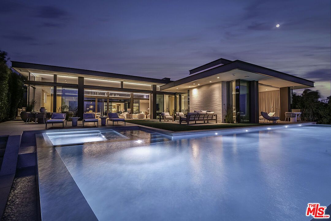 This stunning contemporary estate features a sprawling rear exterior with a seamless indoor-outdoor flow, highlighted by floor-to-ceiling glass walls and a sleek, modern architectural design. A large, illuminated infinity-edge pool and integrated spa serve as the centerpiece of the backyard, perfectly reflecting the twilight sky and the home's warm interior lighting. The expansive patio area is furnished with high-end lounge seating, creating an inviting atmosphere for sophisticated evening entertaining.