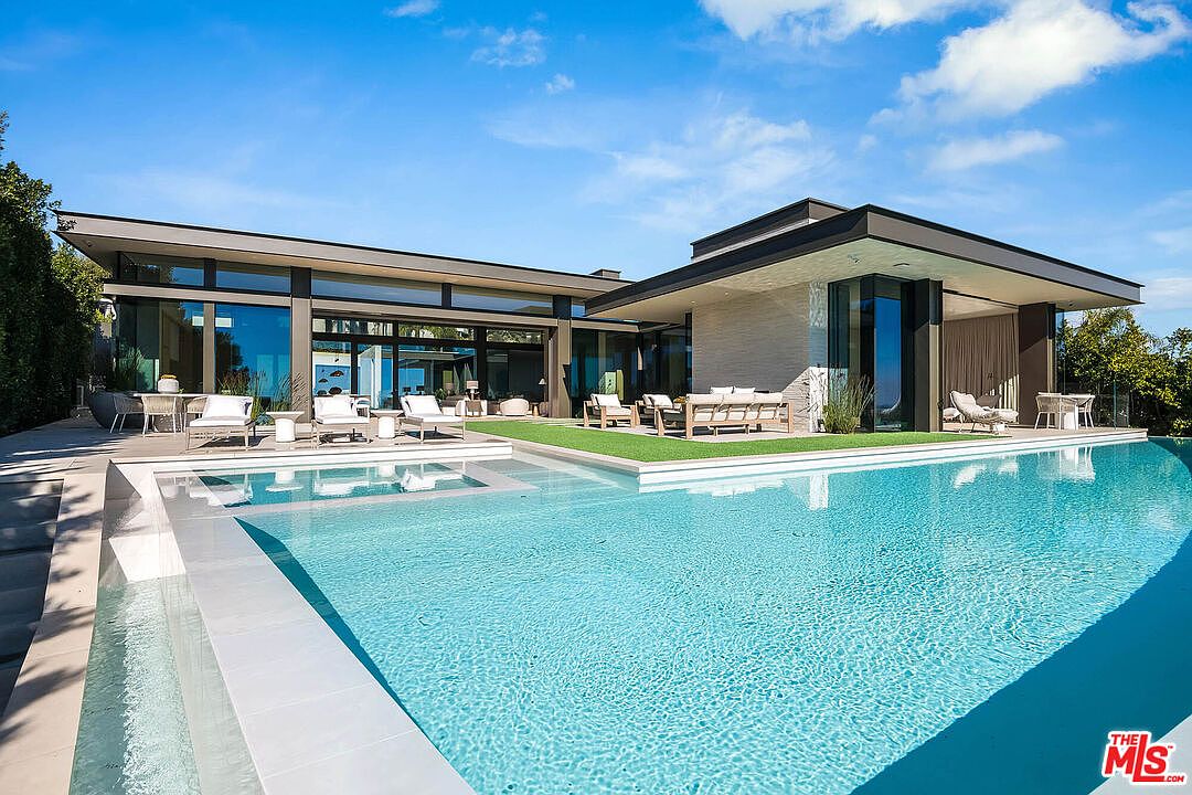 This stunning modern luxury estate features a seamless indoor-outdoor flow with floor-to-ceiling glass walls overlooking a pristine, rectangular swimming pool. The expansive patio area is furnished with contemporary lounge seating, creating an ideal space for high-end entertaining against a backdrop of lush greenery and a clear blue sky. The architectural design emphasizes clean lines, stone accents, and a sophisticated, resort-like atmosphere.
