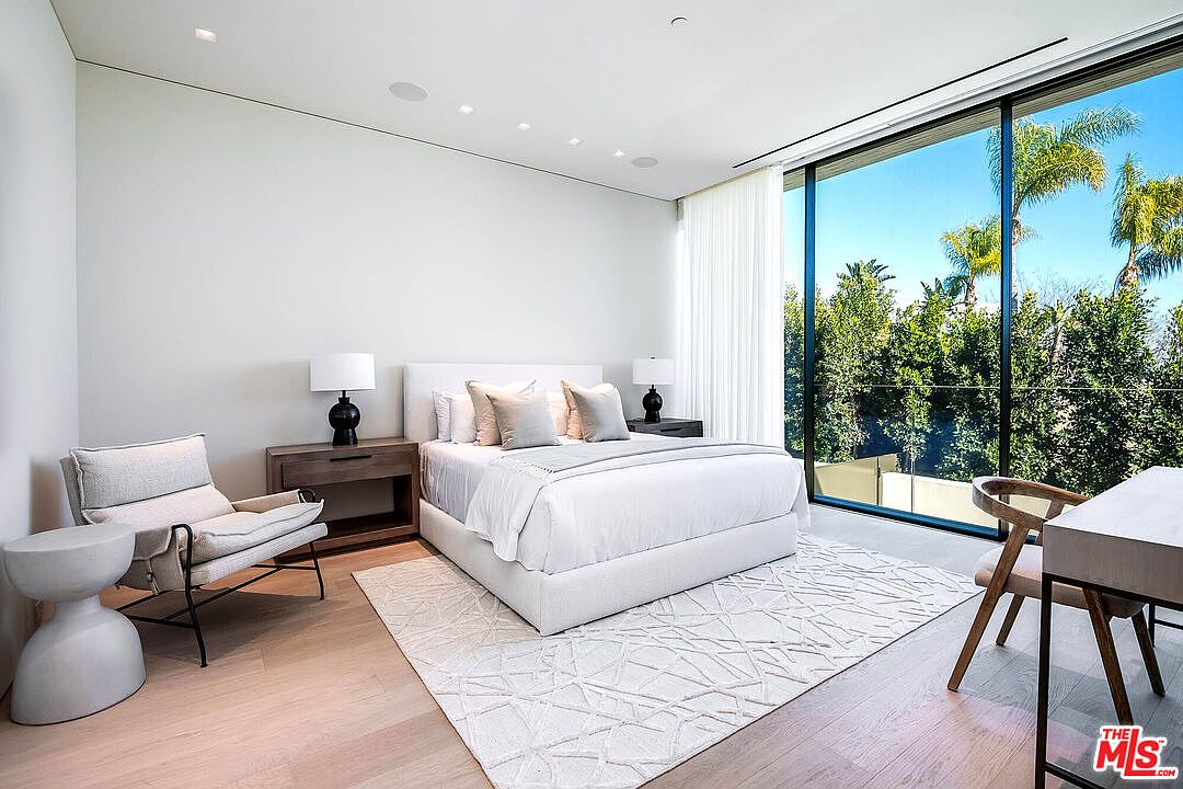 This bright and airy primary bedroom features a minimalist, contemporary design with a plush white bed as the centerpiece, flanked by wooden nightstands and modern lamps. A large floor-to-ceiling window wall offers an expansive view of lush greenery, while a stylish accent chair and side table create a cozy seating area. The room is finished with light hardwood flooring and a textured area rug, evoking a serene and luxurious atmosphere.