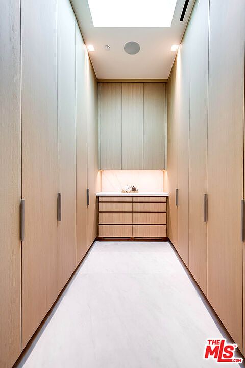 This sophisticated walk-in closet features floor-to-ceiling light wood cabinetry that provides ample storage space, creating a clean and minimalist aesthetic. The central vanity area is illuminated by warm under-cabinet lighting, while a skylight above floods the room with natural light, highlighting the polished marble flooring. The symmetrical design and high-end finishes offer a luxurious and organized atmosphere perfect for a high-end residential property.
