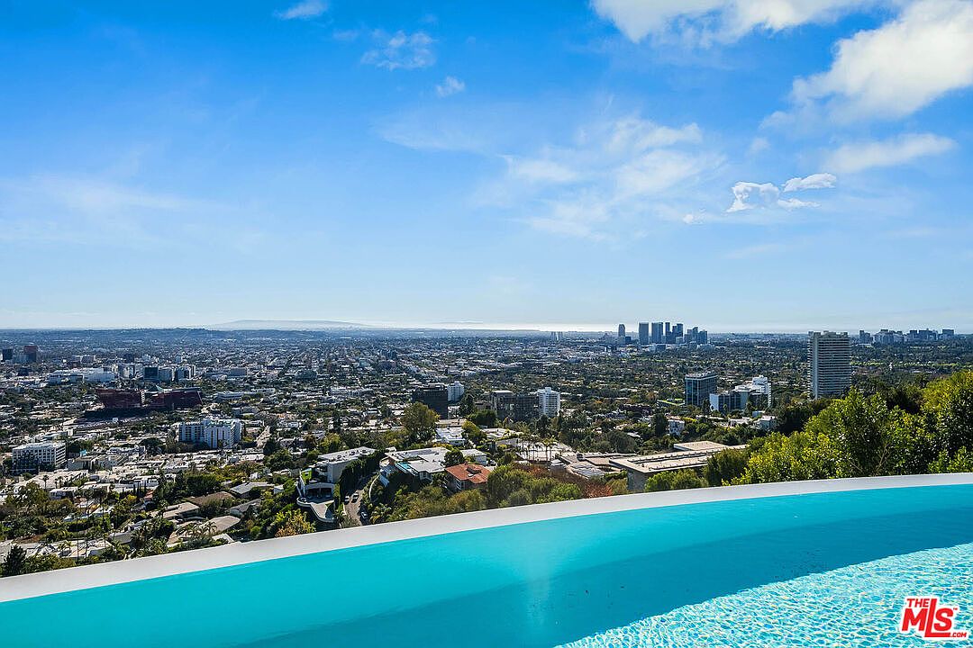 This stunning image captures a breathtaking, elevated view of the Los Angeles skyline from the edge of a luxurious infinity-edge swimming pool. The foreground features the vibrant turquoise water of the pool, which seamlessly blends into the expansive cityscape and distant horizon under a bright, partly cloudy sky. The perspective offers a cinematic sense of grandeur, highlighting the property's prime location and high-end lifestyle appeal.