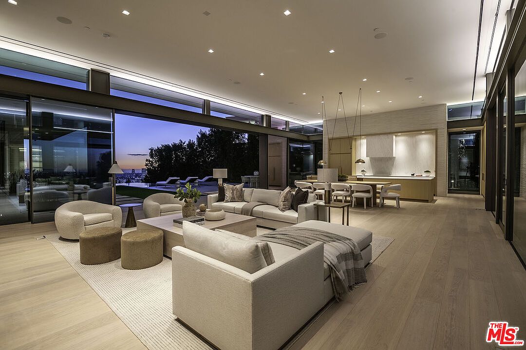 This expansive, open-concept living area showcases a sophisticated modern aesthetic with floor-to-ceiling glass walls that seamlessly blend the interior with a stunning twilight city view. The space is anchored by a plush, neutral-toned sectional and matching armchairs, complemented by a minimalist coffee table and warm wood flooring. The room flows effortlessly into a dining area and kitchen, creating an airy, luxurious atmosphere perfect for high-end entertaining.