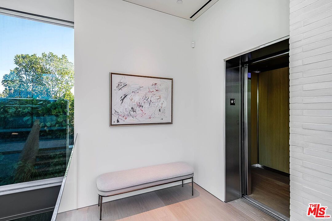 This modern hallway features a sleek, minimalist aesthetic with crisp white walls and light-toned hardwood flooring. A contemporary upholstered bench sits beneath an abstract piece of art, while a polished metal elevator door adds a sophisticated, high-end touch to the space. The perspective captures a bright, airy transition area that seamlessly blends architectural elegance with functional design.