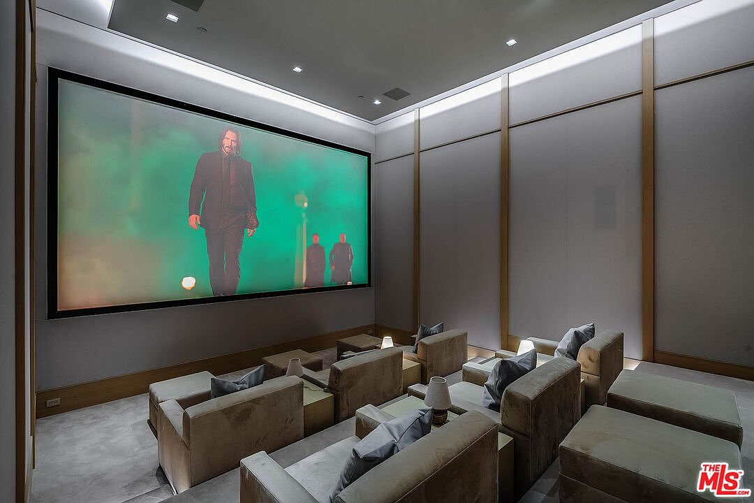 This image showcases a luxurious, tiered home theater room designed for an immersive cinematic experience. The space features multiple rows of plush, neutral-toned armchairs, soft ambient lighting, and a massive projection screen displaying a film. The minimalist aesthetic, characterized by clean lines and sophisticated wall paneling, creates a high-end, professional screening environment.