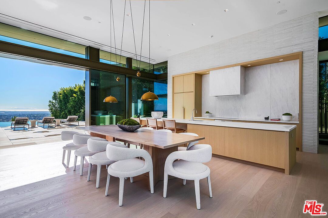 This sophisticated open-concept space seamlessly blends a modern kitchen with a dining area, featuring light wood cabinetry, a minimalist white range hood, and a large island. Floor-to-ceiling glass walls slide open to reveal a stunning outdoor patio and infinity pool overlooking a panoramic city view. The design emphasizes clean lines, natural textures, and a bright, airy atmosphere perfect for luxury indoor-outdoor living.