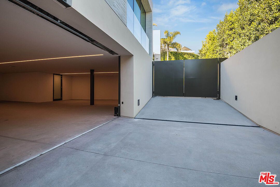 This image showcases a sleek, modern garage area with a wide, open entrance leading into a spacious, minimalist interior. To the right, a large, dark metal gate provides privacy and security, contrasting with the smooth, light-colored concrete driveway and walls. The architectural style is contemporary and clean, emphasizing geometric lines and a sophisticated, high-end aesthetic.