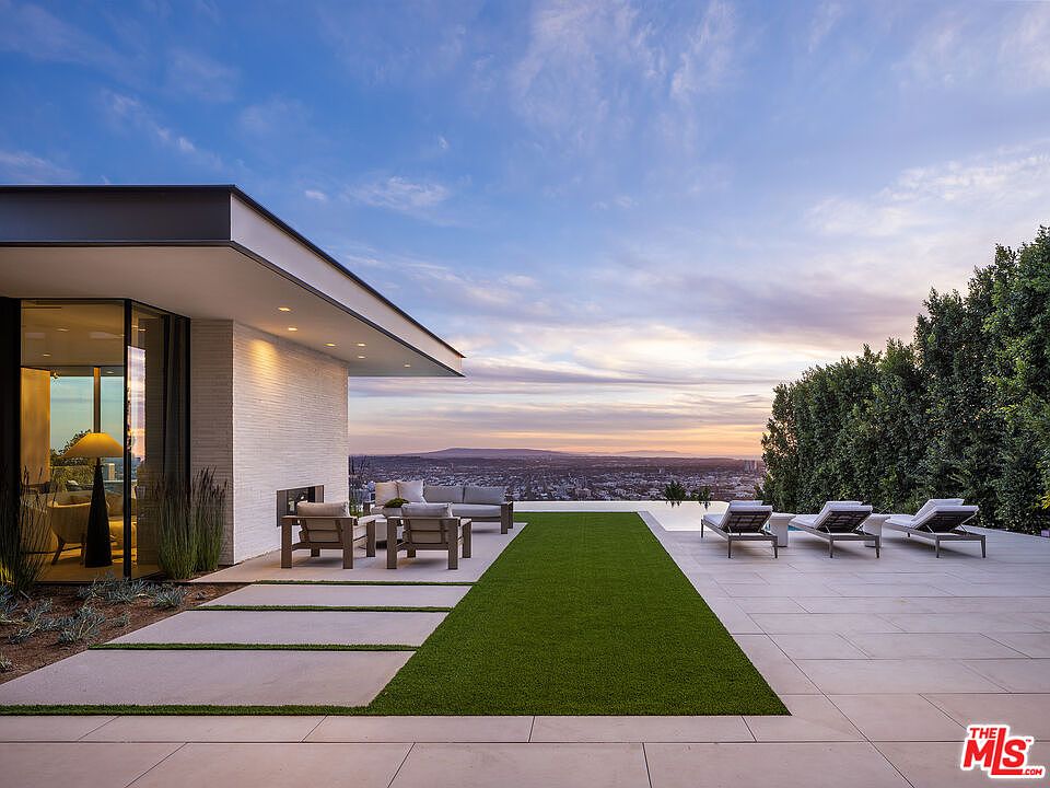 This stunning modern patio features a seamless transition between indoor and outdoor living, highlighted by a sleek infinity-edge pool overlooking a sprawling city skyline at sunset. The space is furnished with contemporary lounge seating and sun loungers, set against a backdrop of manicured greenery and clean architectural lines. The perspective captures the expansive, luxurious atmosphere of a high-end hillside estate.