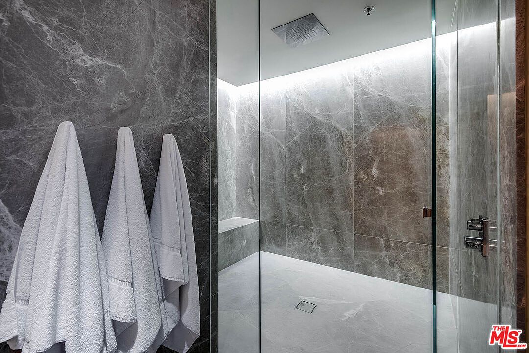 This luxurious walk-in shower features floor-to-ceiling dark grey marble tiling and a sleek, frameless glass enclosure. A built-in stone bench and a modern rainfall showerhead enhance the spa-like atmosphere, while soft, recessed LED lighting adds a sophisticated glow. Three plush white towels hang conveniently on the adjacent wall, emphasizing the high-end, contemporary design of the space.