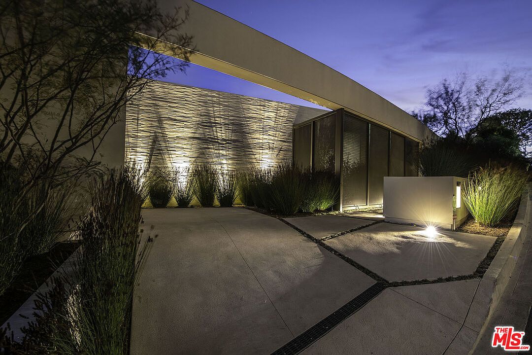 This striking modern entryway features a dramatic, cantilevered architectural beam framing a textured stone wall illuminated by warm uplighting. The scene is captured at dusk, highlighting the clean lines of the concrete driveway and the minimalist landscaping that creates a sophisticated, high-end aesthetic. The perspective is low and wide, emphasizing the grand scale and contemporary design of the home's facade.