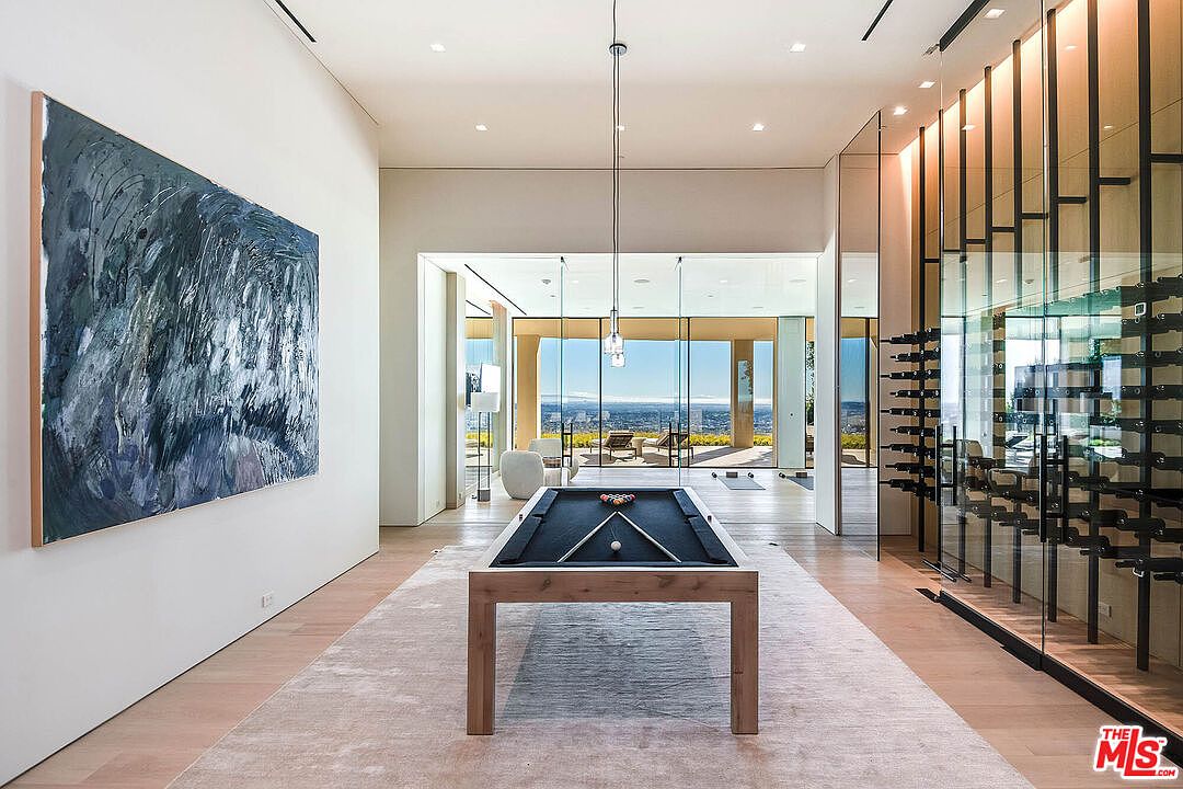 This sophisticated interior space features a minimalist design centered around a sleek pool table, set against a backdrop of floor-to-ceiling glass walls that reveal a stunning panoramic view. To the left, a large, abstract blue-toned painting adds a bold artistic statement, while a glass-enclosed wine cellar on the right provides a luxurious, functional display. The room is bathed in natural light, emphasizing the clean lines, light wood flooring, and high-end contemporary aesthetic.