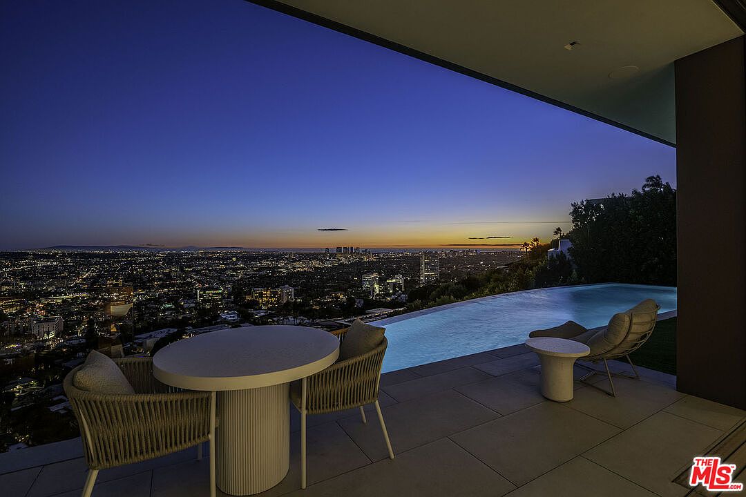 This stunning outdoor terrace offers a breathtaking panoramic view of the city skyline at dusk, framed by a sleek, modern architectural overhang. The space features a minimalist dining set and a lounge chair positioned beside a luxurious infinity-edge pool that appears to merge with the horizon. The atmosphere is sophisticated and serene, highlighting the property's premium location and high-end design.