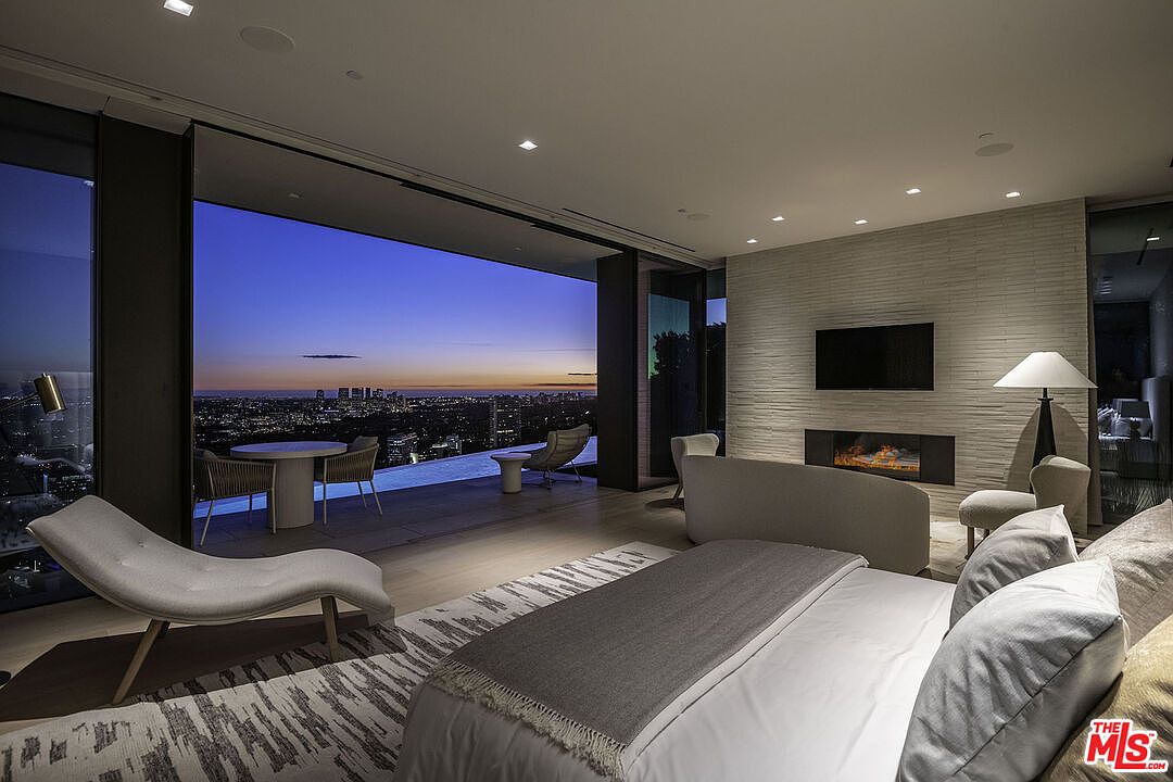 This luxurious primary bedroom features a contemporary design with a seamless transition to an expansive outdoor balcony overlooking a stunning city skyline at dusk. The room is anchored by a plush bed and a sleek fireplace set into a textured stone wall, creating a sophisticated and tranquil atmosphere. The open-concept layout and floor-to-ceiling glass walls emphasize the breathtaking panoramic views, highlighting the property's high-end architectural appeal.