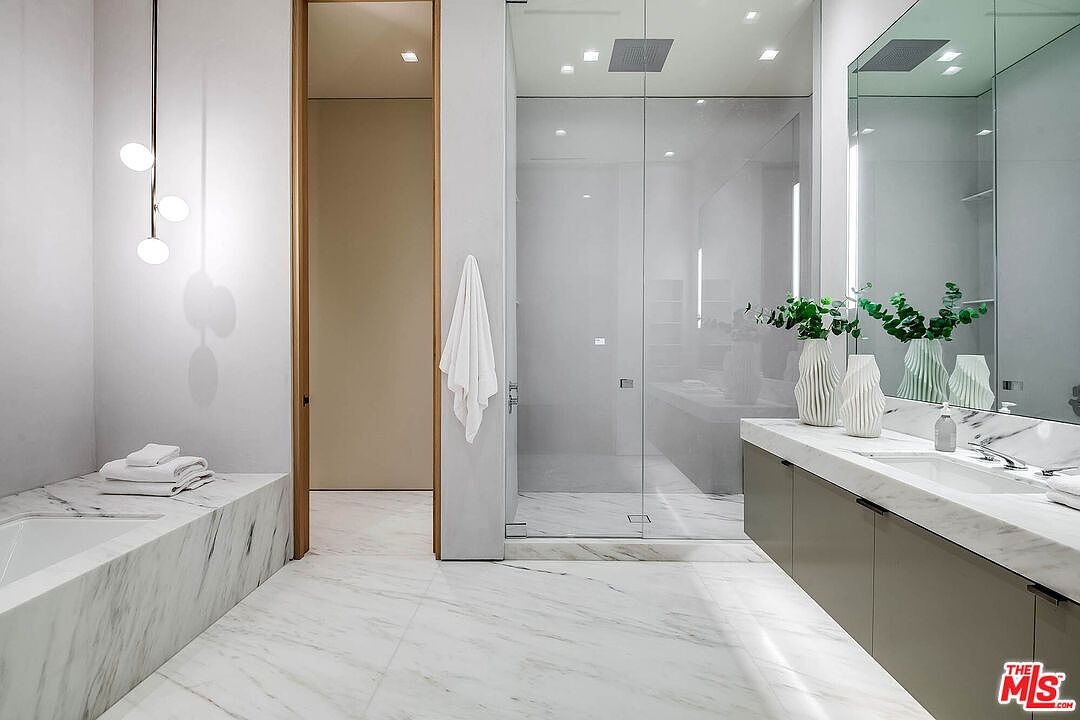 This luxurious primary bathroom features a sleek, modern aesthetic with extensive use of white marble on the floors and the bathtub surround. The space includes a double vanity with a marble countertop, a large glass-enclosed walk-in shower, and elegant pendant lighting hanging over the tub area. The neutral color palette and clean lines create a sophisticated, spa-like atmosphere.