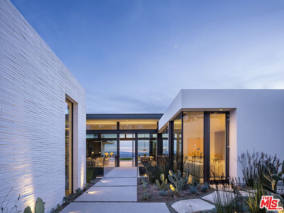 This striking modern entryway features a clean, minimalist aesthetic with a combination of white brick and smooth stucco walls. A concrete paver walkway leads through a desert-inspired landscape toward a glass-walled corridor that offers a transparent view through the home to the horizon beyond. The scene is captured at dusk, with warm interior lighting contrasting against the cool blue twilight sky, creating an inviting and sophisticated atmosphere.