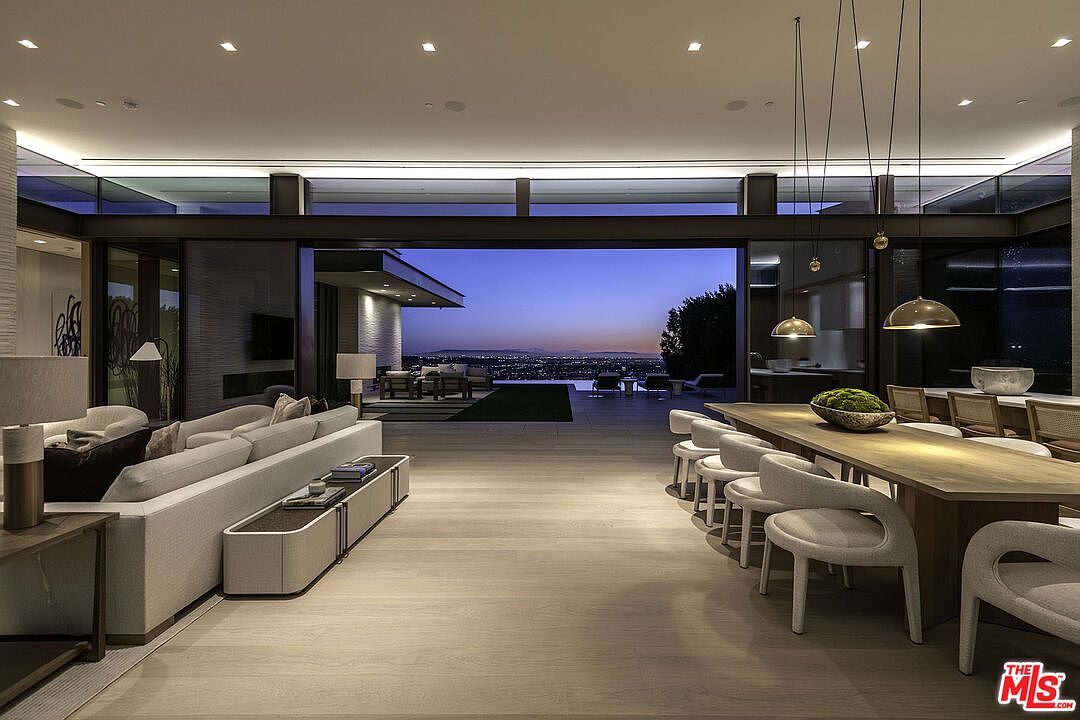 This expansive, open-concept living and dining area features a seamless transition to an outdoor patio through floor-to-ceiling glass walls, showcasing a breathtaking panoramic city view at dusk. The interior is defined by a sophisticated, modern aesthetic with neutral-toned furnishings, light wood flooring, and elegant pendant lighting over the long dining table. The perspective captures the harmonious blend of luxury indoor living and the dramatic, illuminated cityscape beyond.