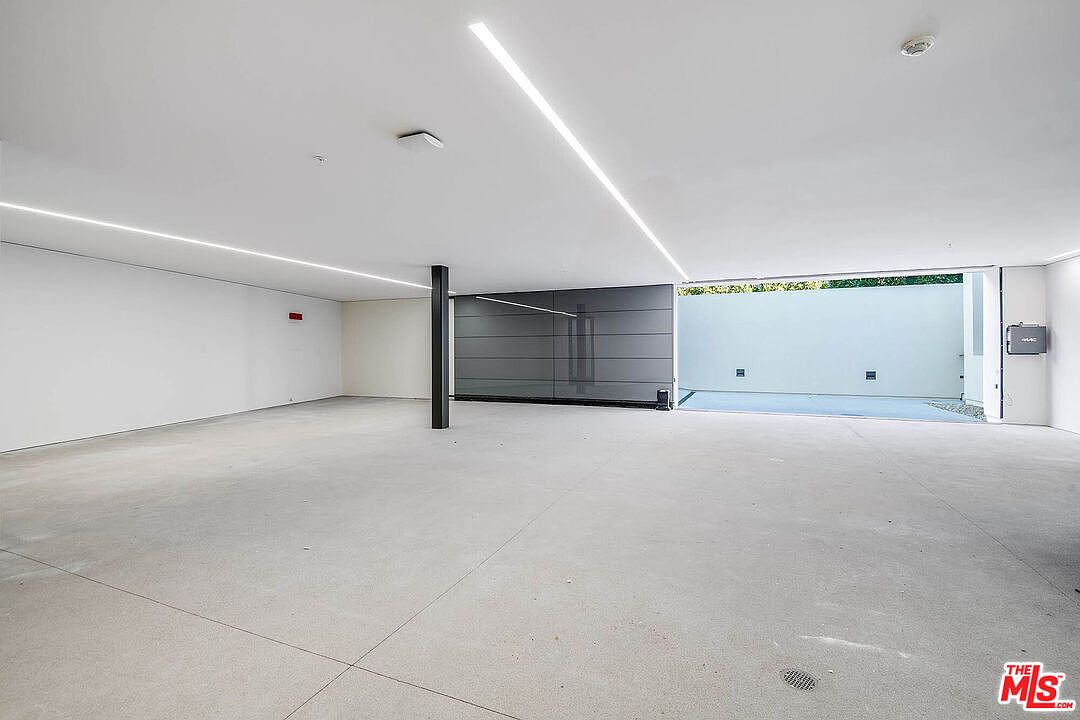 This spacious, modern garage features a minimalist design with smooth concrete flooring and clean, white walls illuminated by recessed linear lighting. The open-concept layout provides ample room for multiple vehicles, while a large, dark-paneled door leads to an outdoor area, creating a seamless transition between indoor and outdoor spaces. The overall aesthetic is sleek, contemporary, and highly functional.