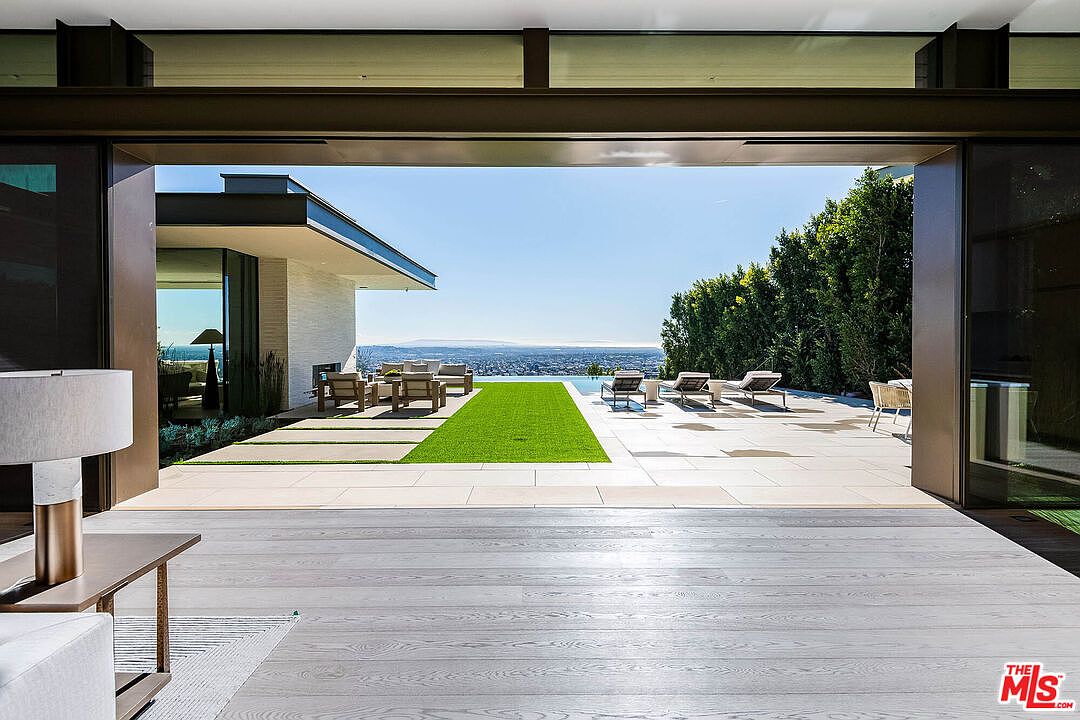 This image captures a seamless transition from a modern interior living space to an expansive outdoor patio and pool deck through massive, open sliding glass doors. The outdoor area features a manicured green lawn strip, comfortable lounge seating, and a stunning infinity-edge pool that overlooks a sprawling city view under a clear blue sky. The architectural design emphasizes indoor-outdoor living with clean lines, high-end materials, and a sophisticated, luxurious aesthetic.