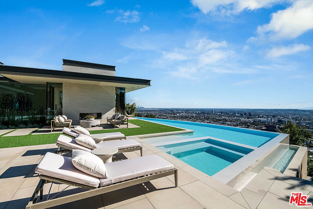 This stunning modern luxury estate features a seamless infinity-edge pool and integrated spa overlooking a sprawling panoramic city view. The expansive stone patio is furnished with sleek lounge chairs and a cozy outdoor seating area, creating an ideal space for high-end entertaining. The architecture emphasizes clean lines and a sophisticated indoor-outdoor flow, perfectly capturing the essence of upscale hillside living.