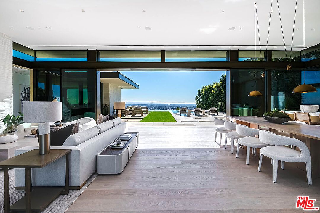 This expansive, open-concept living and dining area features a seamless transition to the outdoors through massive floor-to-ceiling glass walls that frame a stunning panoramic view. The interior is defined by a sophisticated, minimalist aesthetic with light hardwood floors, a plush grey sectional sofa, and a long wooden dining table illuminated by modern pendant lights. The perspective captures the airy, luxurious atmosphere of a high-end modern residence, emphasizing the integration of indoor and outdoor living spaces.